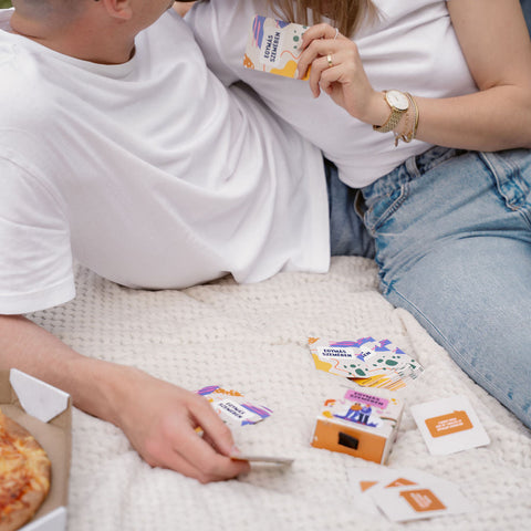 Fiatal pár fehér takarón ül, párkapcsolati kártyajátékkal játszanak, mellettük pizza látható